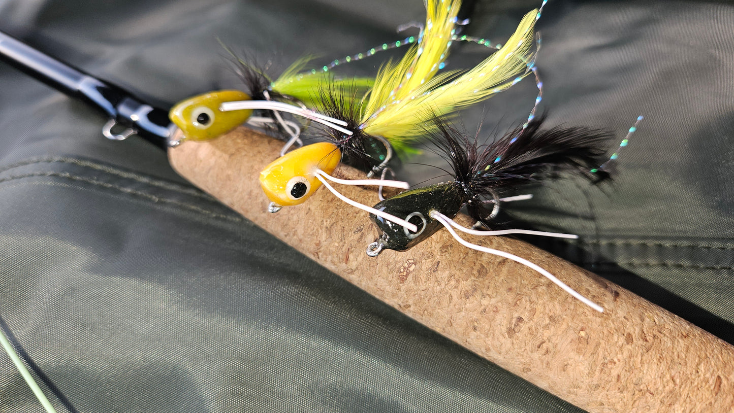 Three fishing flies tied on a cork rod, with one yellow fly featuring black and green feathers and the other a yellow body with black hackle and white hackle wings, set against a dark olive green fabric background.